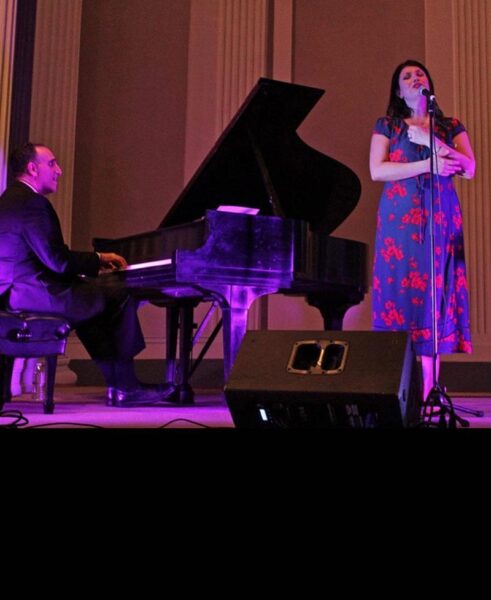 A man in a suit plays a grand piano while a woman in a floral dress sings into a microphone on a stage with purple lighting.