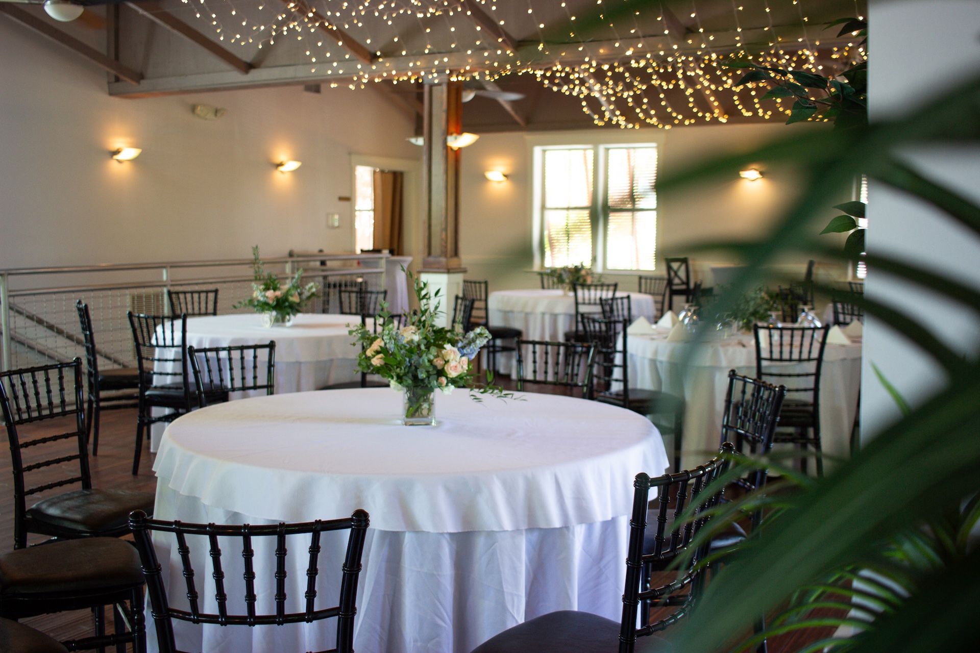 A bright event space with round tables covered in white tablecloths, black chairs, floral centerpieces, and string lights hanging from the ceiling. Sunlight streams through windows, and green plants are visible in the foreground.