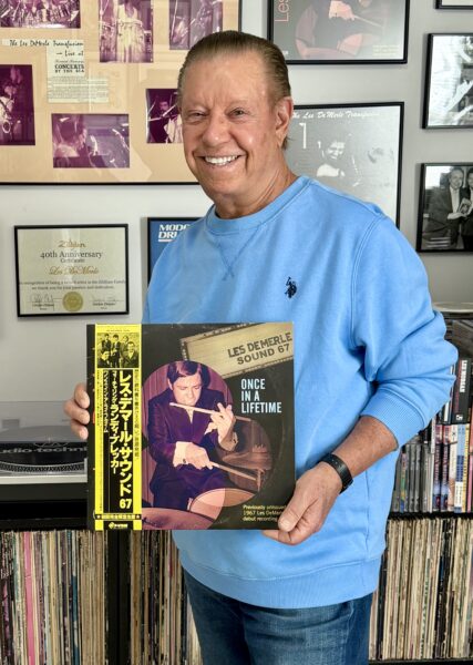 A smiling man in a blue sweatshirt stands in front of a wall with framed records and photos, holding a vinyl record titled Once in a Lifetime by Les Demerle Sound 67. Shelves of records line the background.