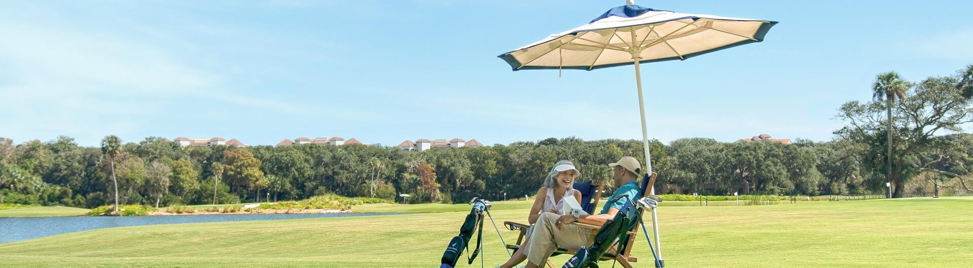 Two people sit on chairs under a large umbrella on a golf course, with golf bags beside them. Trees and a lake are in the background under a clear blue sky.