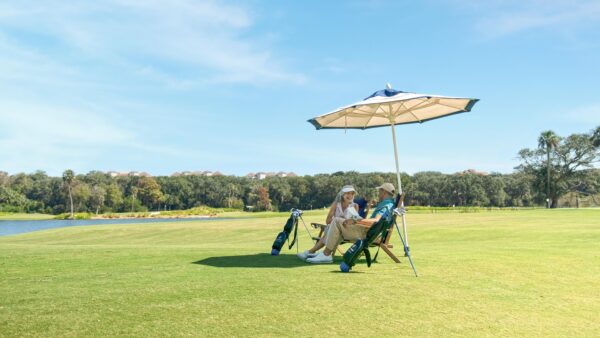 Two people sit on chairs under a large umbrella on a golf course, with golf bags beside them. Trees and a lake are in the background under a clear blue sky.