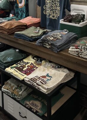 A display table in a store with neatly folded graphic t-shirts, some with nature and animal designs. More shirts are stored on the lower shelf and drawers underneath, all on a wooden floor with a patterned rug.