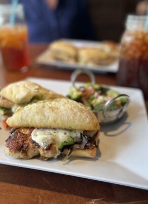 A close-up of a sandwich with grilled chicken, melted cheese, and greens on ciabatta bread, served on a square white plate with a small salad and a drink in the background.