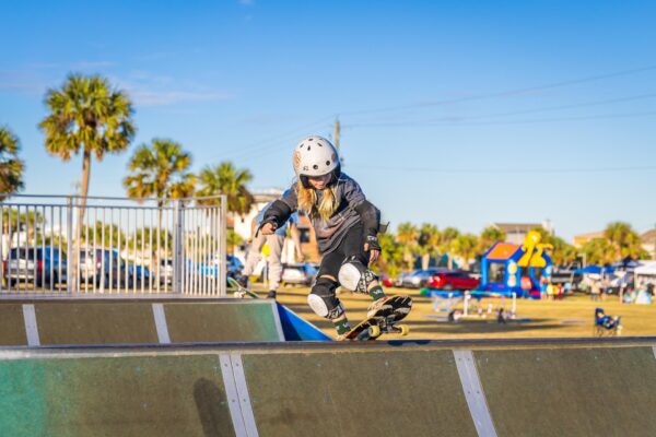 A young child wearing a helmet and protective gear skateboards on a ramp at an outdoor skate park, with palm trees, cars, and people in the background under a clear blue sky.