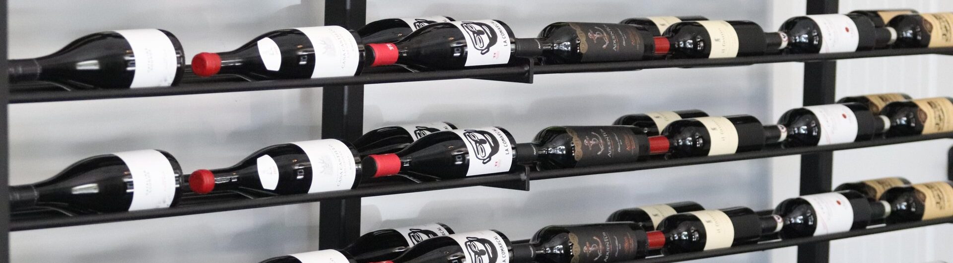Rows of wine bottles with various labels and red foil tops are neatly arranged on a black metal rack against a white wall. The bottles are stored horizontally in a modern, organized display.