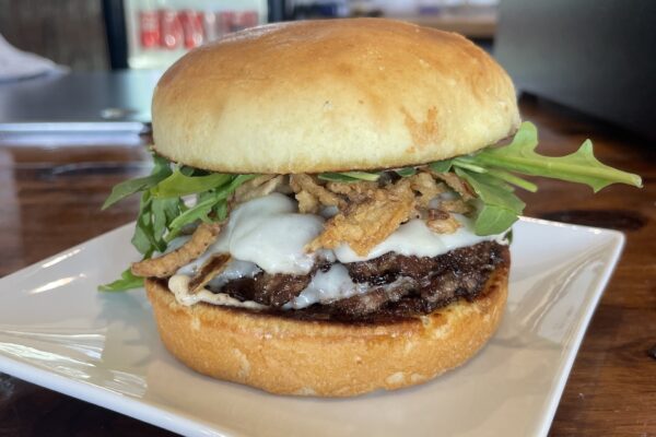 A close-up of a gourmet burger with melted cheese, crispy fried onions, arugula, and a beef patty on a toasted bun, served on a square white plate.