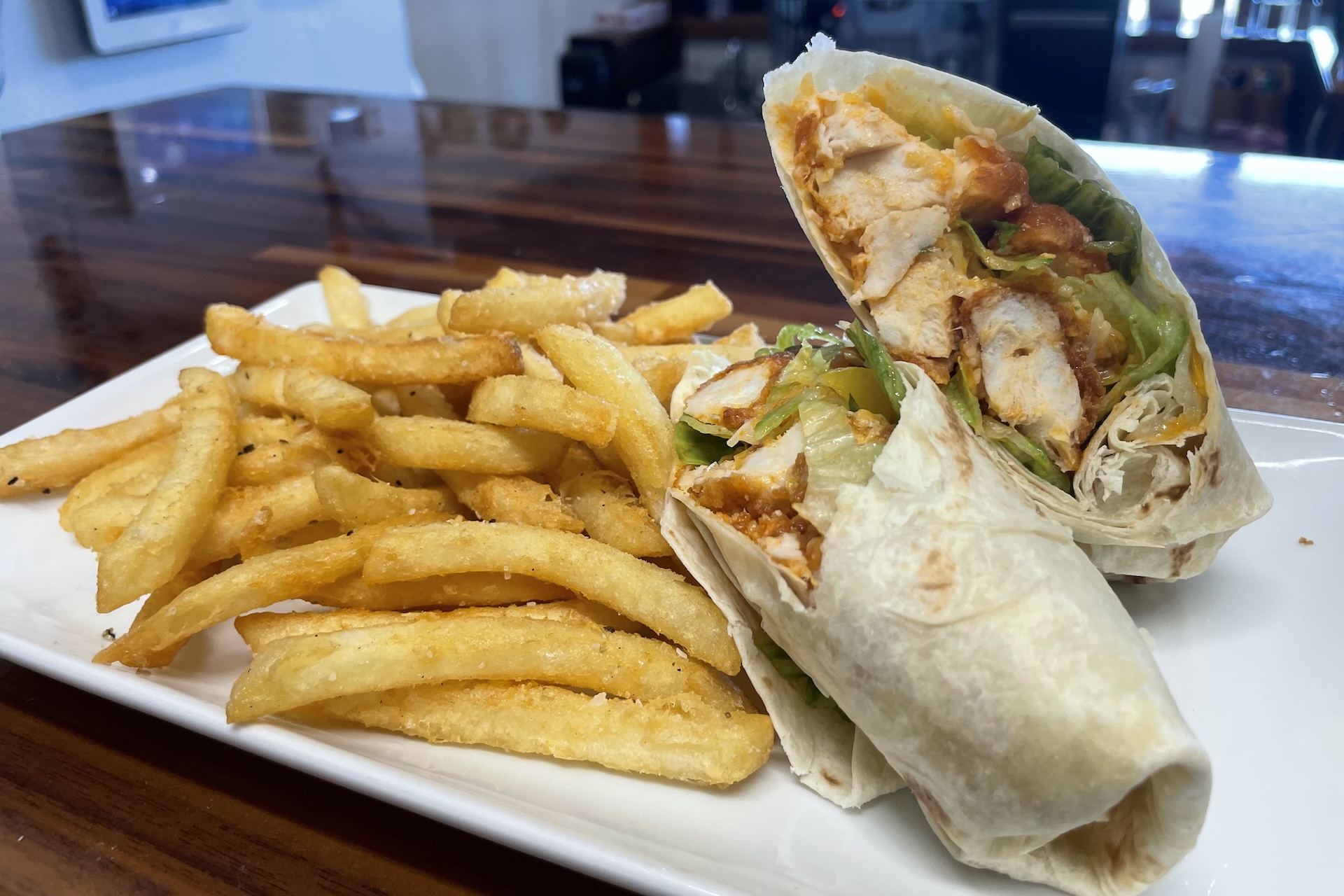 A plate of golden French fries next to a sliced chicken wrap filled with lettuce, tomato, and grilled chicken, served on a white rectangular plate on a wooden table.