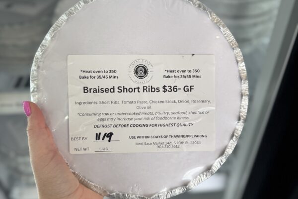 A hand holds a frozen packaged meal labeled Braised Short Ribs $36 - GF. The label lists ingredients, heating instructions, and a best by date of 11/9. The package is round, covered with white paper and silver foil.
