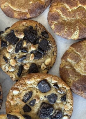 A tray of freshly baked cookies: some topped with crushed Oreos and white chocolate chips, and others coated with cinnamon sugar, likely snickerdoodles, all arranged on parchment paper.