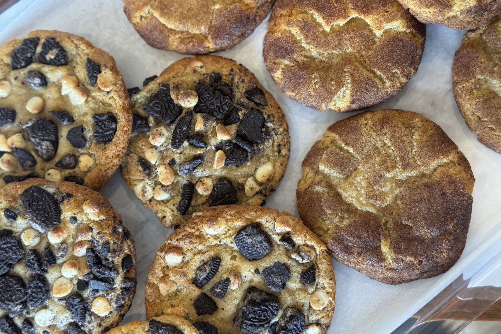 A tray of freshly baked cookies: some topped with crushed Oreos and white chocolate chips, and others coated with cinnamon sugar, likely snickerdoodles, all arranged on parchment paper.