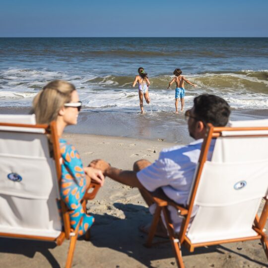 A man and woman sit in beach chairs facing the ocean, holding hands, while two children play at the edge of the water under a clear blue sky.
