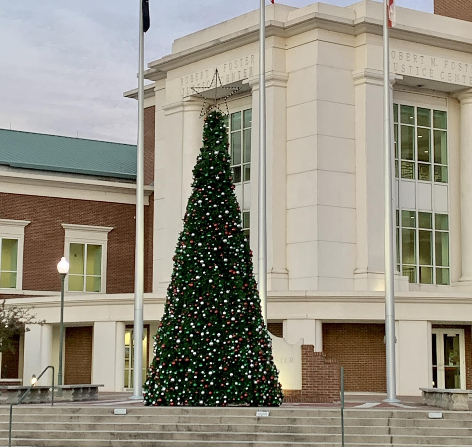 A large decorated Christmas tree with a star on top stands in front of a building with columns and large windows. Three flagpoles are in front of the tree, and steps lead up to the area.