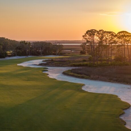 A scenic golf course at sunset with a green fairway bordered by white sand bunkers, trees on the left, and open marshland on the right under a glowing orange sky.