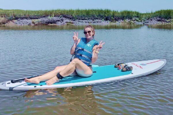 A woman in a blue life vest sits smiling on a paddleboard in calm water, making peace signs with both hands. Grassy banks and blue sky are visible in the background.
