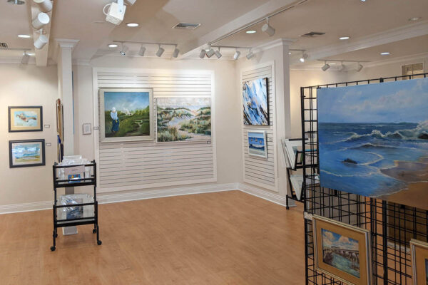An art gallery with landscape paintings on white walls and a black display grid, featuring seascapes, dunes, and a person painting outdoors. The room has wood floors and overhead track lighting.
