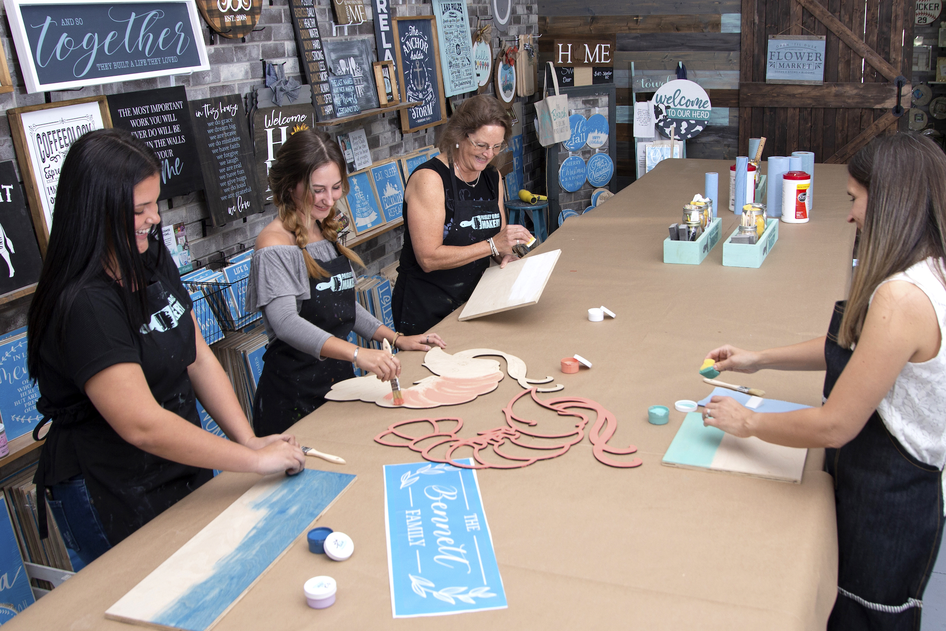 Four women in a craft studio smile while painting wooden signs and cutouts on a large table. The workspace is decorated with finished signs, paint supplies, and wall art.