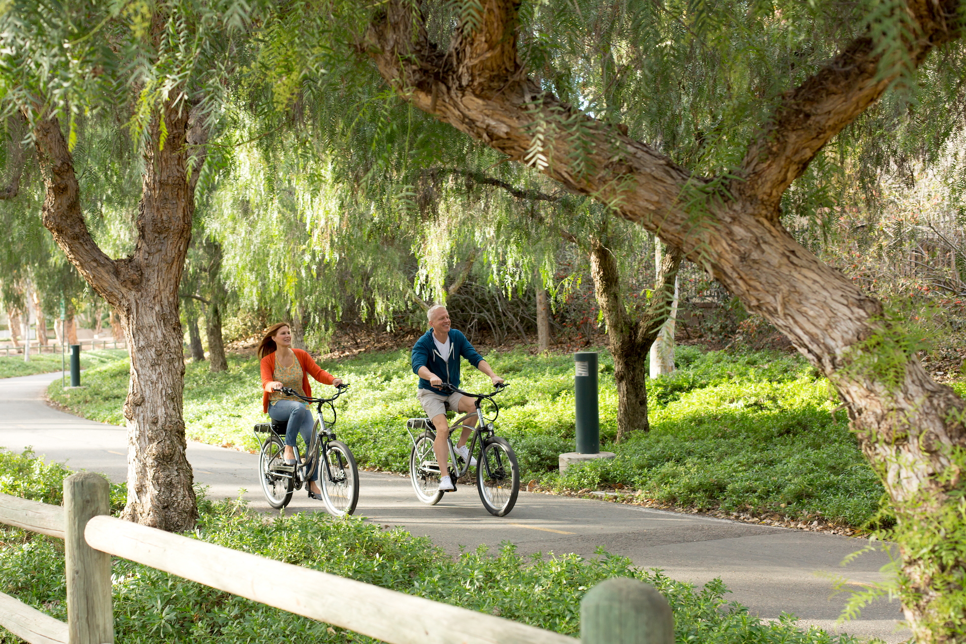 A woman and a man ride bicycles along a winding paved path surrounded by green trees and lush plants on a sunny day. A wooden fence runs alongside the trail. Both appear relaxed and are enjoying the outdoors.
