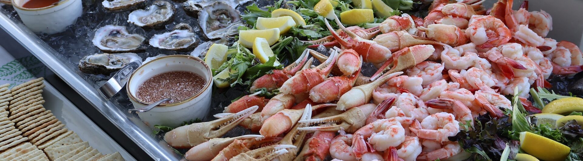 A seafood platter with oysters, crab legs, and shrimp on ice, garnished with lemon wedges and leafy greens. Dipping sauces and a plate of crackers are placed on the side.