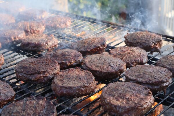 Rows of beef patties cooking on a grill with smoke rising and flames visible beneath, suggesting an outdoor barbecue setting.
