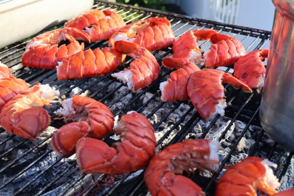 Lobster tails are grilling on an outdoor barbecue, their shells bright red and meat exposed. Sunlight highlights the seafood, and there are ashes and a metal pot nearby on the grill.