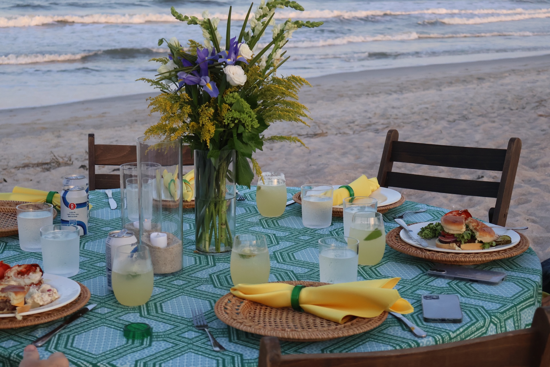 A round table set with plates of food, drinks, and yellow napkins sits on the sand by the ocean. A vase of flowers and candles decorate the table. Waves are visible in the background, creating a relaxed beachside atmosphere.