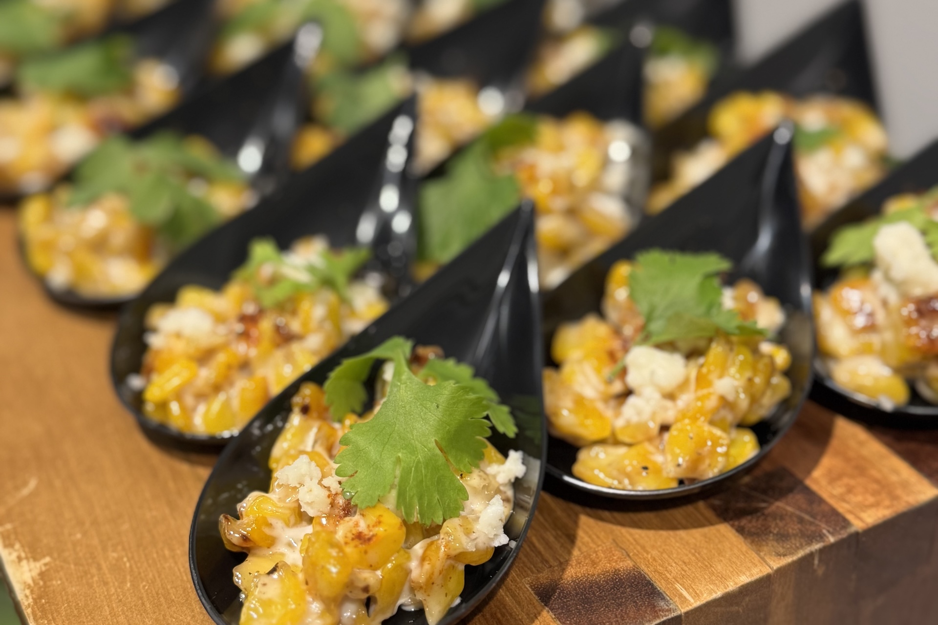 Black appetizer spoons filled with a creamy yellow corn mixture, garnished with crumbled cheese and cilantro, are arranged in rows on a wooden surface.