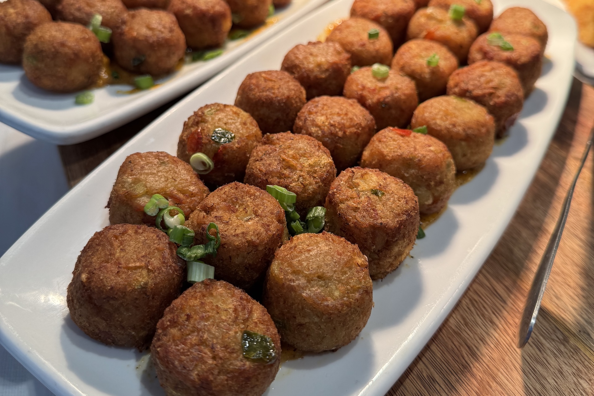 Rows of golden-brown fried balls are neatly arranged on white rectangular plates, garnished with chopped green onions, and set on a wooden table.