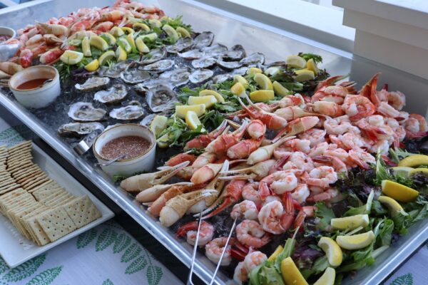 A seafood platter with oysters, crab legs, and shrimp on ice, garnished with lemon wedges and leafy greens. Dipping sauces and a plate of crackers are placed on the side.