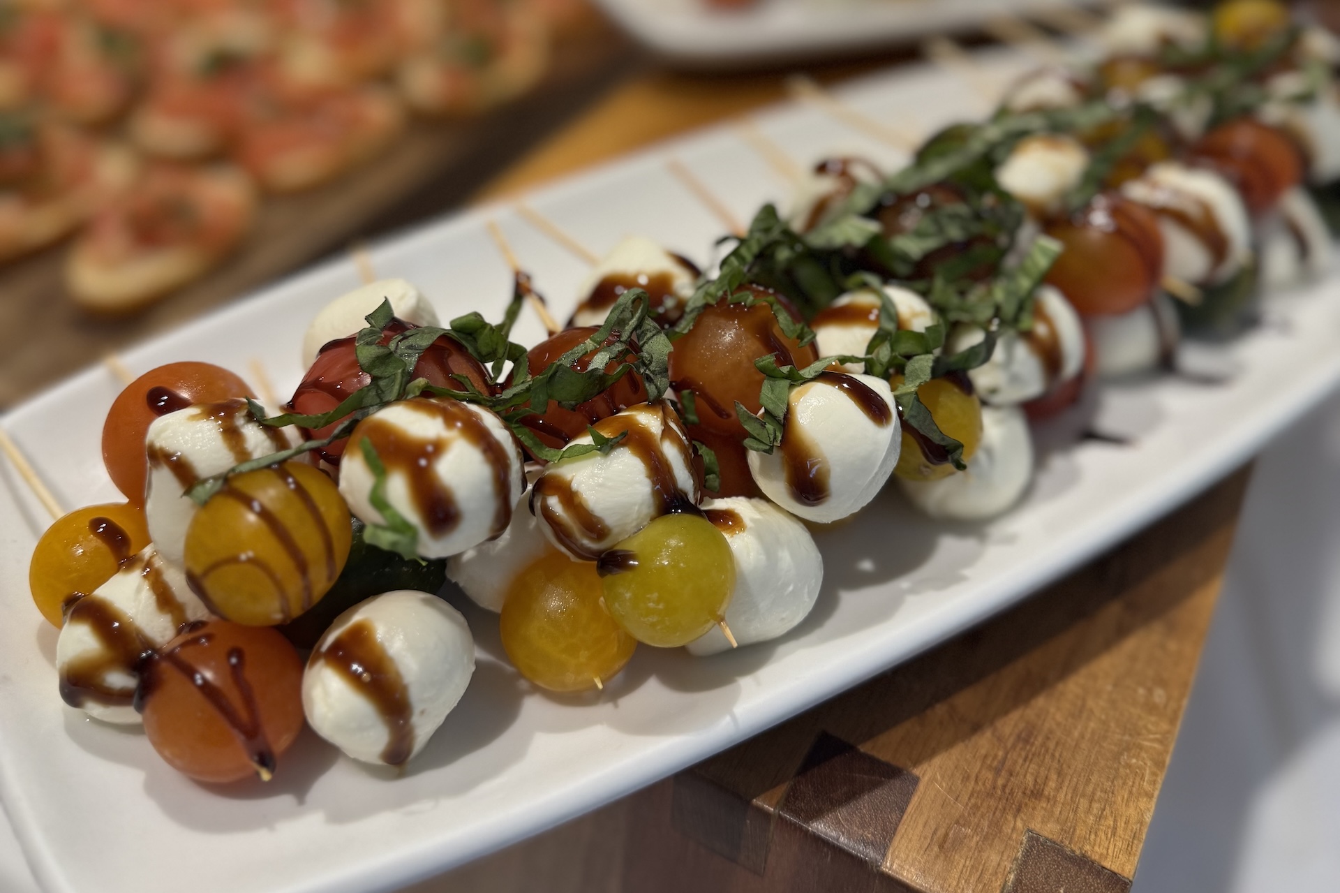 Skewers of cherry tomatoes, fresh mozzarella balls, and basil leaves drizzled with balsamic glaze are arranged on a white platter. In the background, there are more appetizers on a wooden board.