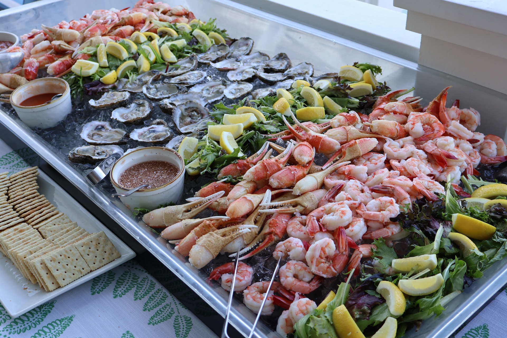 A seafood platter with oysters, crab legs, and shrimp on ice, garnished with lemon wedges and leafy greens. Dipping sauces and a plate of crackers are placed on the side.