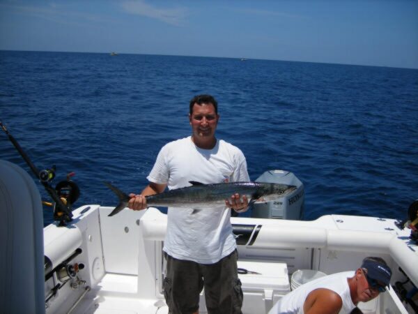 A man standing on a boat holds a large fish, smiling at the camera. The boat is on open water under a clear blue sky, with another person seated nearby and fishing equipment visible.