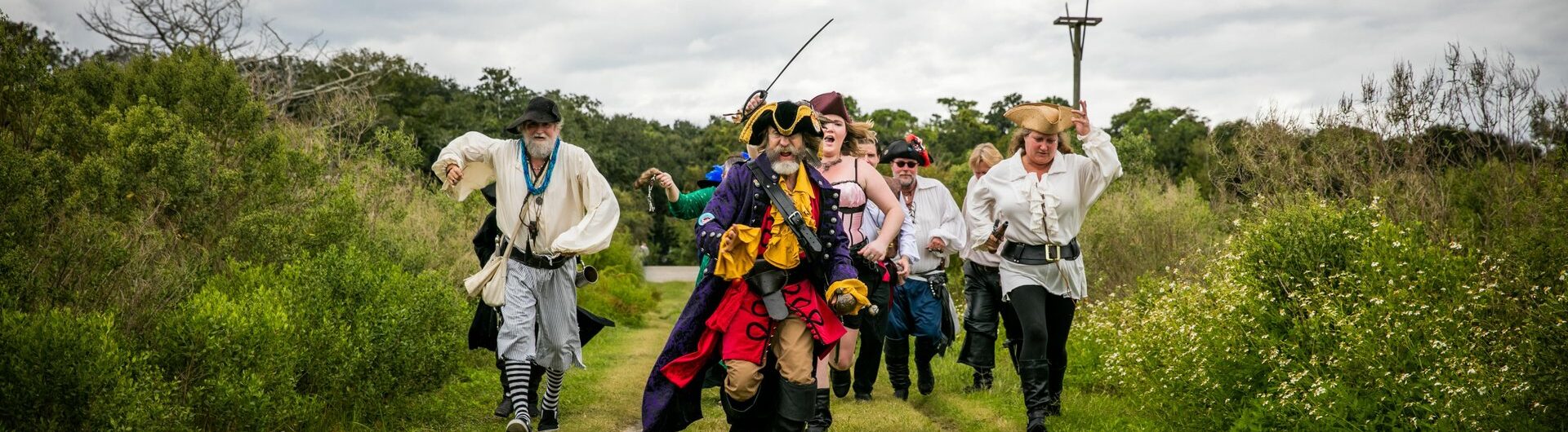 A group of people dressed as pirates, led by a person in a colorful pirate costume with a hat and sword, walk energetically down a grassy path surrounded by greenery under a cloudy sky.