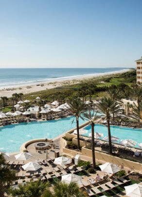 Aerial view of a beachfront resort with palm trees, multiple swimming pools, lounge chairs, umbrellas, and a large hotel building beside a sandy beach and the ocean under a clear blue sky.