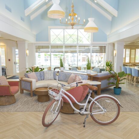A bright, spacious lounge with pastel furniture, indoor plants, wooden tables, and a white bicycle with a basket in the center. Large windows let in natural light and a chandelier hangs from the vaulted ceiling.