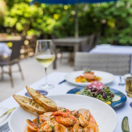 A bowl of seafood stew with shrimp, clams, and bread slices sits on a white tablecloth, accompanied by a glass of white wine. Out-of-focus outdoor seating and a blue umbrella are visible in the background.