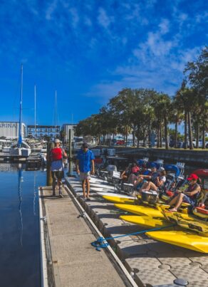 People prepare yellow water bikes at a dock beside moored boats at Amelia Island Marina, with trees and blue sky in the background. Some people stand while others sit on the water bikes.