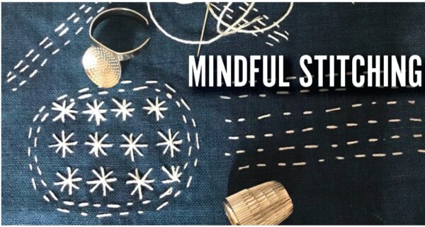 A navy blue fabric with white stitched patterns, a needle with thread, a thimble, and a metal ring. Bold text on the fabric reads “MINDFUL STITCHING.”.