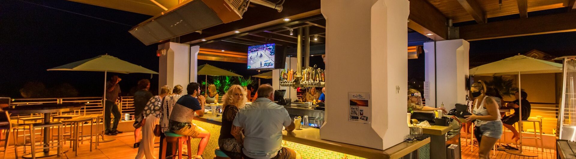 A rooftop bar at night with people sitting on stools around the bar, chatting and watching a TV. Warm yellow lighting and wooden décor create a relaxed atmosphere. Staff are serving drinks and people are enjoying themselves.