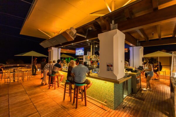 A rooftop bar at night with people sitting on stools around the bar, chatting and watching a TV. Warm yellow lighting and wooden décor create a relaxed atmosphere. Staff are serving drinks and people are enjoying themselves.