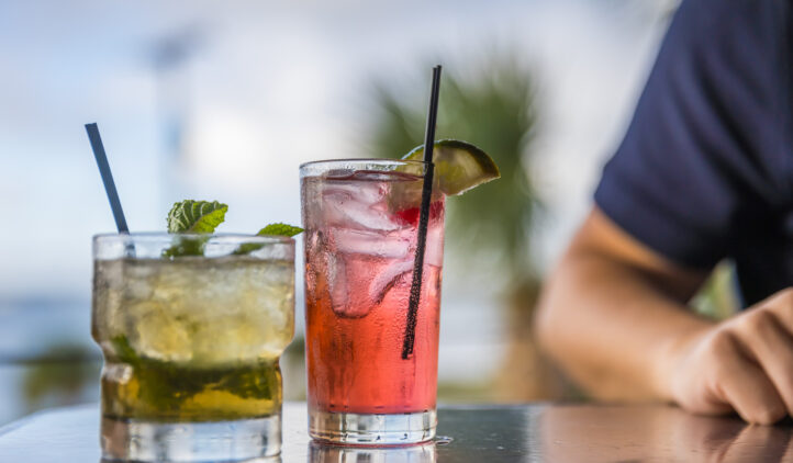 Two colorful cocktails on a reflective table: one greenish drink with mint, and one pink drink with a lime and cherry. Both have straws. A person with a navy sleeve sits in the blurred background with palm trees visible.
