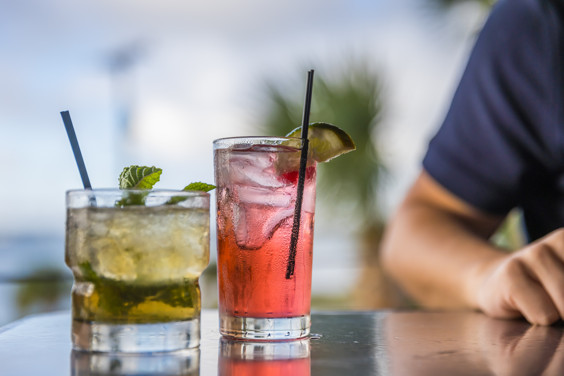 Two colorful cocktails on a reflective table: one greenish drink with mint, and one pink drink with a lime and cherry. Both have straws. A person with a navy sleeve sits in the blurred background with palm trees visible.