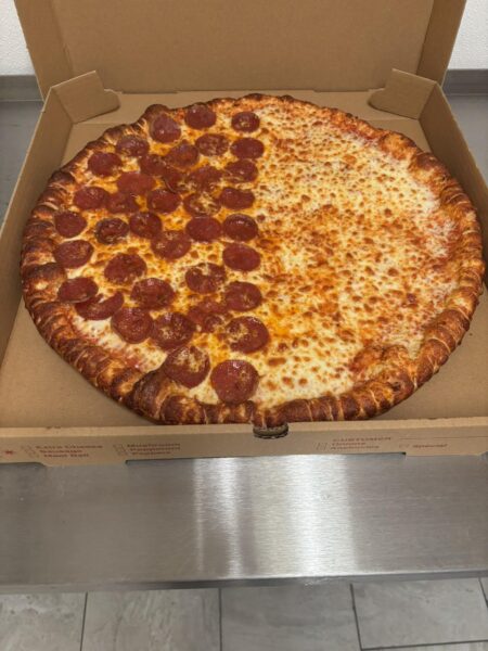 A large pizza in an open cardboard box sits on a metal counter. Half the pizza is topped with pepperoni slices, while the other half is plain cheese.