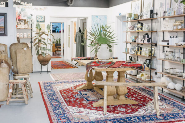 A bright boutique features wooden furniture, colorful patterned rugs, shelves with decorative items, potted plants, and framed artwork displayed on white walls. A large green plant in a vase sits on a central table.
