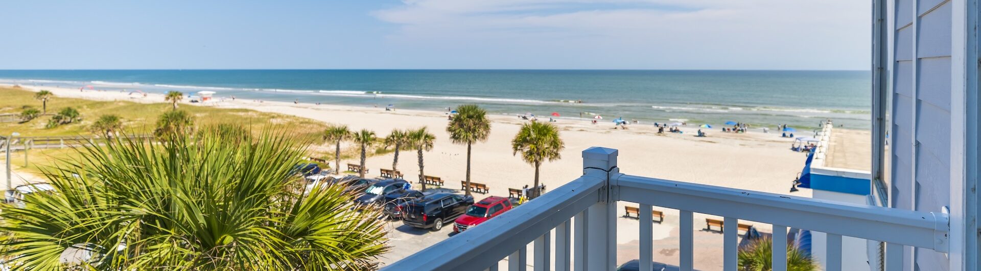 View from a balcony overlooking a sunny beach with white sand, palm trees, parked cars, and people relaxing near the ocean. A white chair and railing are visible in the foreground. Blue sky with a few clouds overhead.