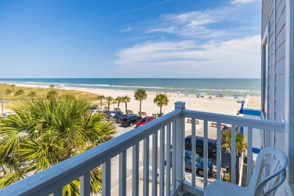 View from a balcony overlooking a sunny beach with white sand, palm trees, parked cars, and people relaxing near the ocean. A white chair and railing are visible in the foreground. Blue sky with a few clouds overhead.