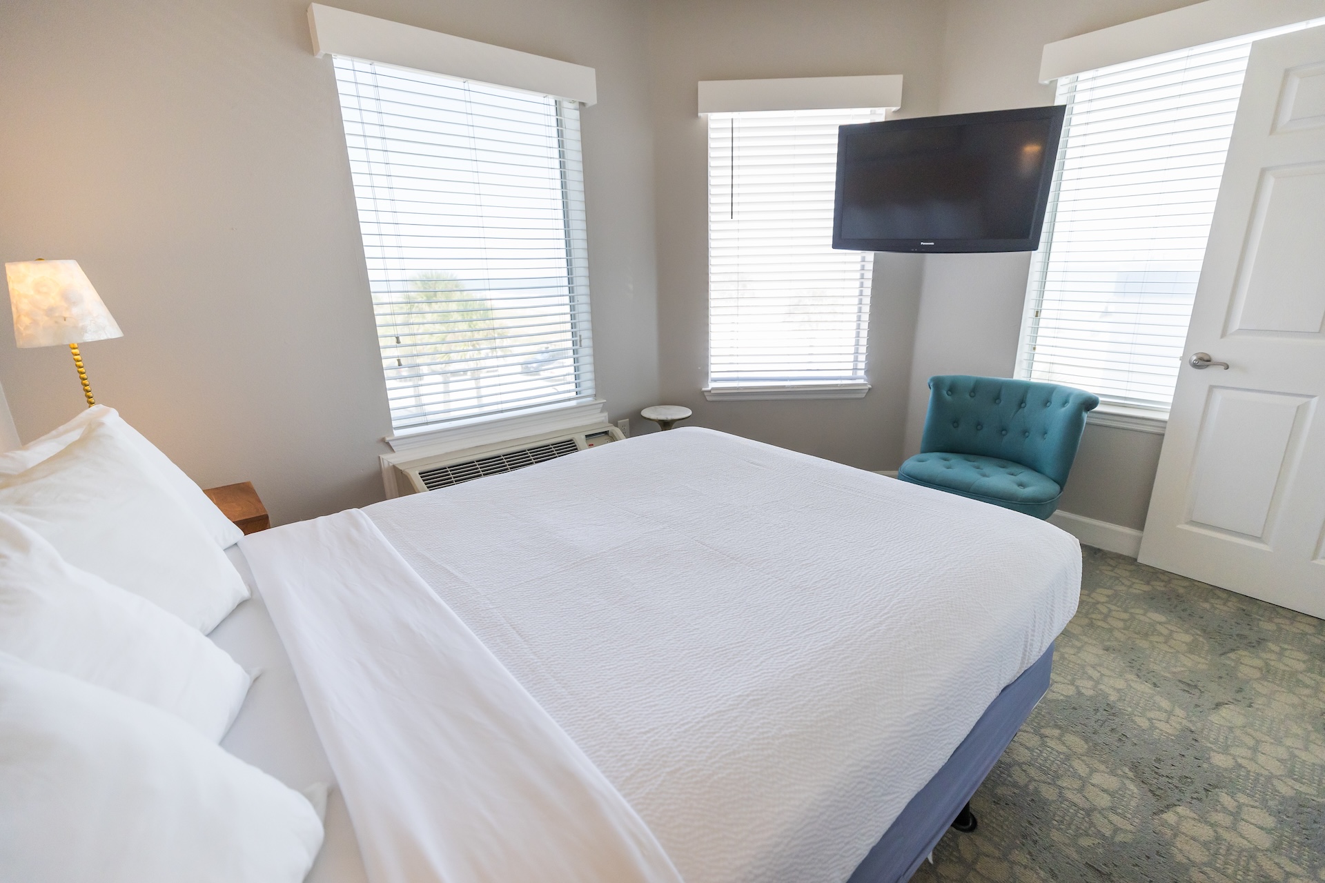 A bright bedroom with a neatly made bed, white bedding, a blue tufted chair, wall-mounted TV, lamp, air conditioner, and large windows with blinds letting in natural light.