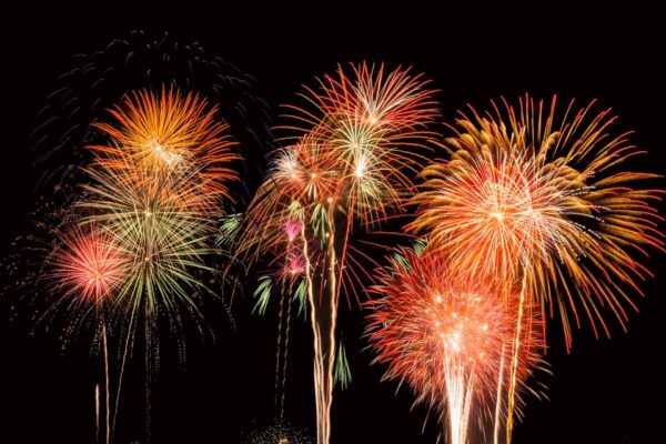 Colorful fireworks burst in the night sky, displaying vibrant red, orange, yellow, and green patterns against a black background. Several fireworks are exploding simultaneously, creating a festive atmosphere.