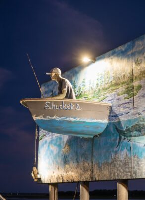 A boat labeled “Shuckers” with a mannequin fisherman is mounted on a mural-covered wall, illuminated by a spotlight at night, with a twilight sky in the background.