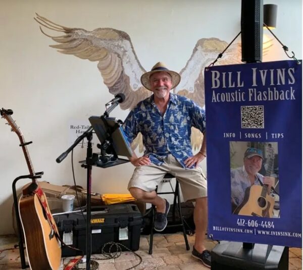 A smiling man in a straw hat and blue shirt sits on a stool with musical equipment and guitars, with painted wings on the wall behind him. A banner reads Bill Ivins Acoustic Flashback with contact info and a photo.