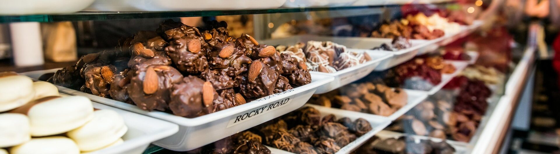 A display case filled with various types of chocolate candies and confections on white trays, labeled with names like “Oreo” and “Rocky Road,” in a brightly lit candy shop.